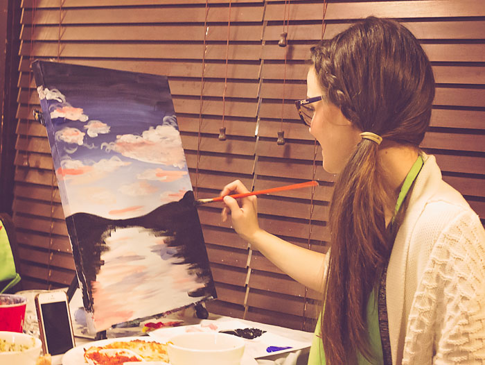 A woman painting a canvas. The canvas has a dark blue faded sky reflecting onto a river, which is surrounded by black silhouetted trees.