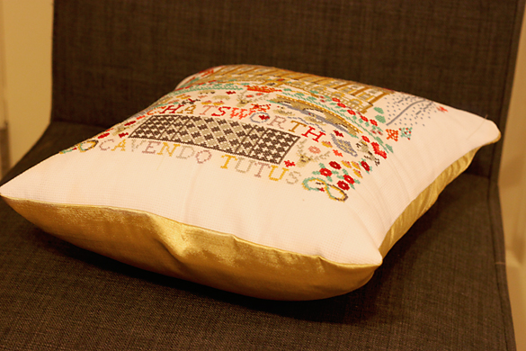 A cross-stitched pillow of different elements related to Chatsworth House in Chesterfield, England, laying flat on a chair.