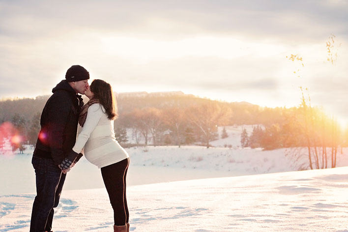 A man and a pregnant woman facing each other in the foreground, with a wintery river valley blurred in the background.