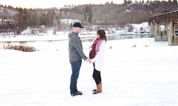 A husband wearing a baseball cap standing facing his wife, who is wearing a burgundy flowy shirt over her 8-month pregnant belly.