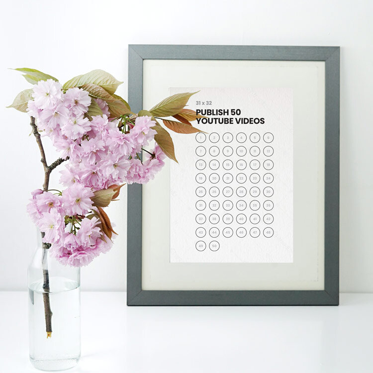 A picture frame with a 50-day challenge tracker printed; there is a bold title and 50 bubbles with tiny numbers inside to fill in and track your progress. There is also a pink flower in a glass vase.