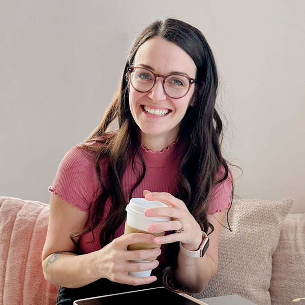 Bex has long brown curly hair, big glasses, and an even bigger smile. She is sitting in a coffee shop, and has a white to-go coffee cup in her hand.